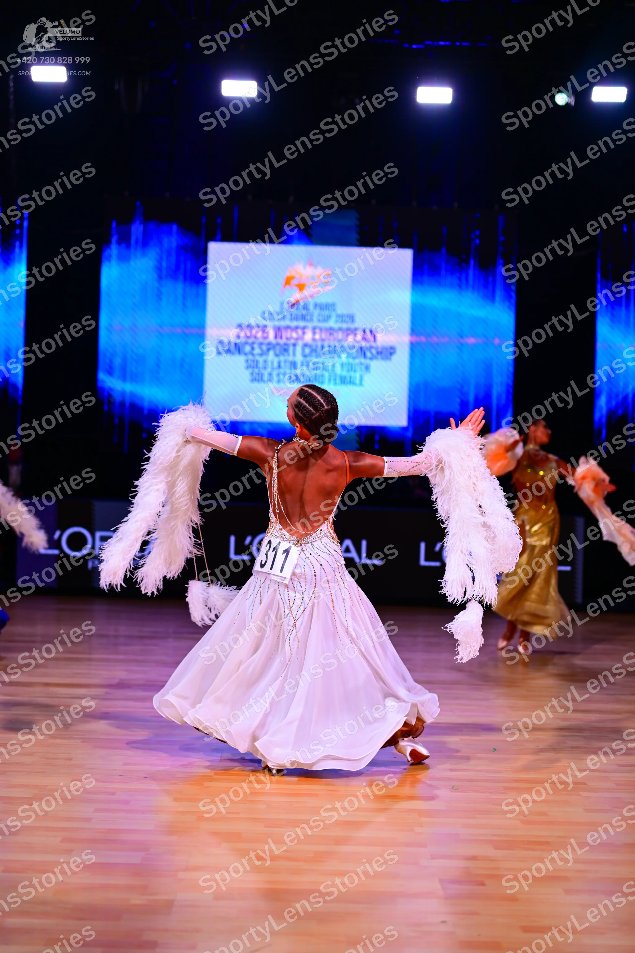 Dancesport competitor: Female dancer in foreground: white dress with large white feathery arm accessori