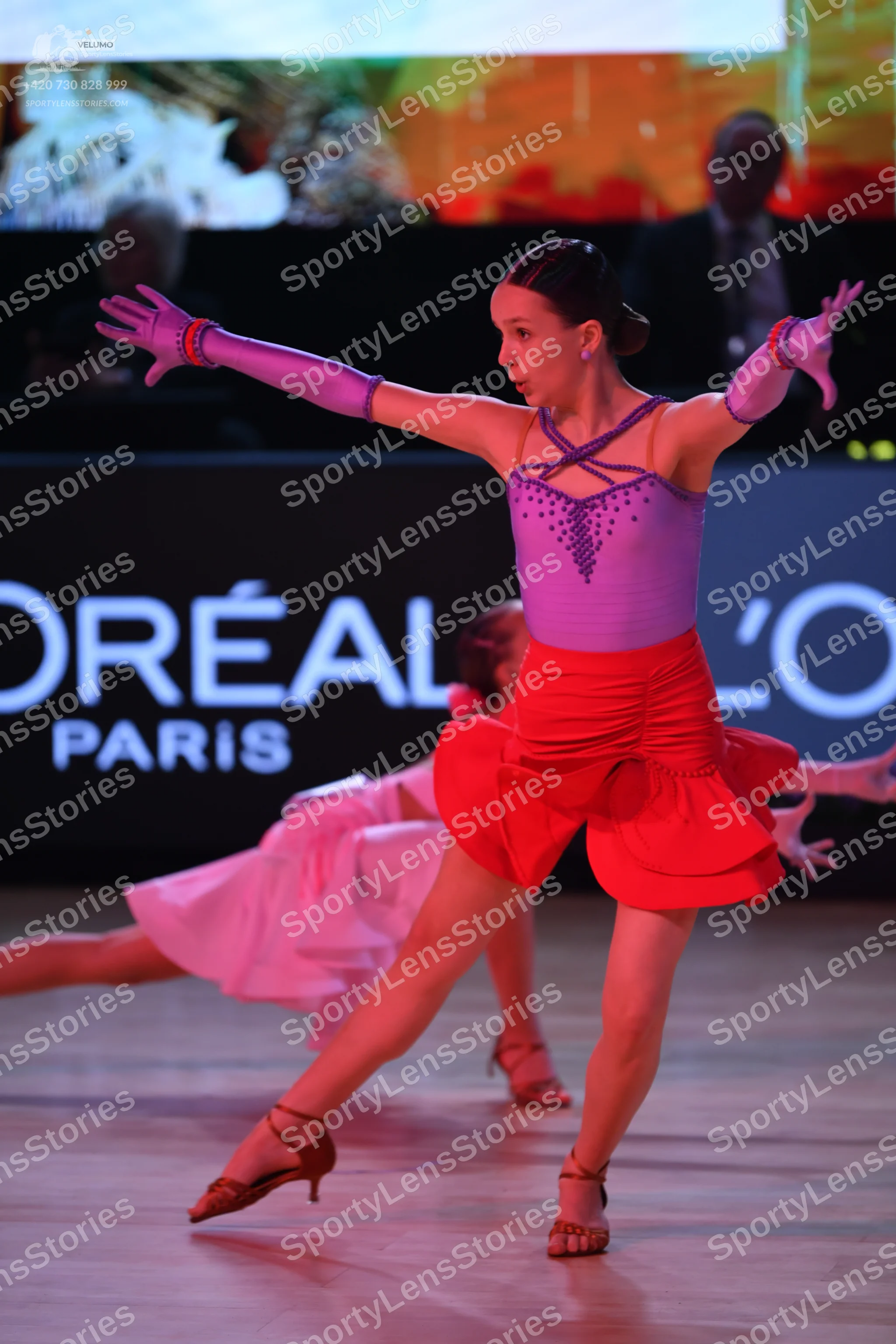 Dancesport competitor: Female dancer in foreground: lilac bodice with sequin patterns, red ruffled skir