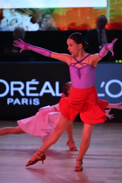 Dancesport competitor: Female dancer in foreground: lilac bodice with sequin patterns, red ruffled skir