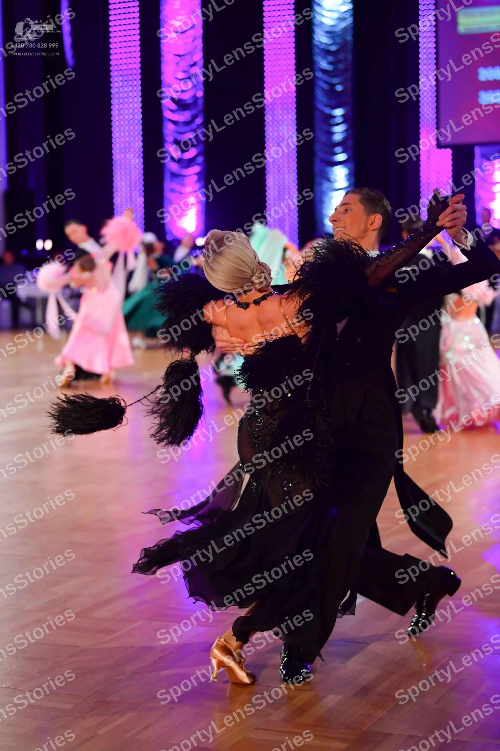 Dancesport competitor: Foreground female dancer: black ballroom dress with large black feather-like fri
