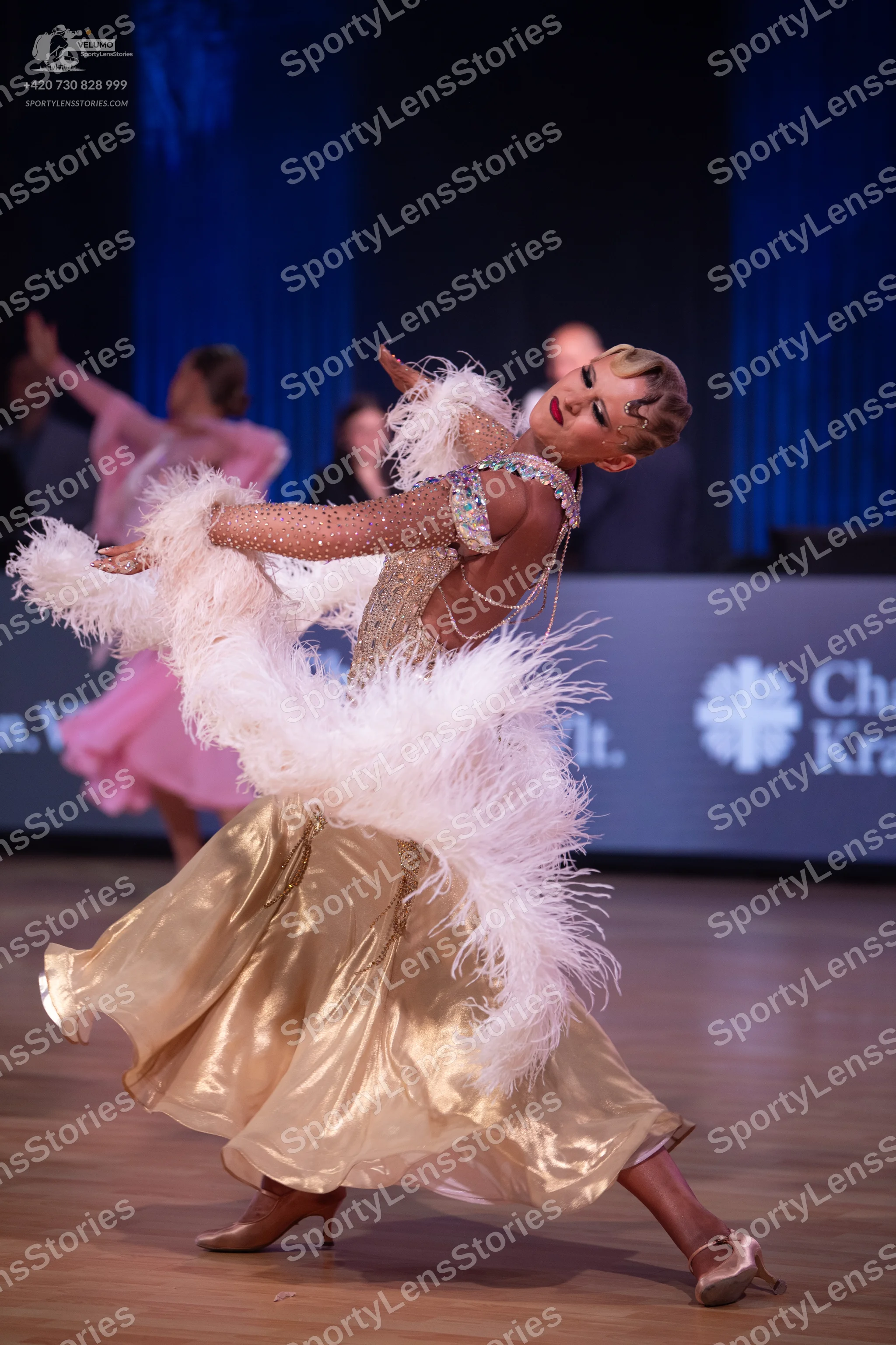 Dancesport competitor: Female dancer in foreground: gold ballgown with a sequined bodice and large whit