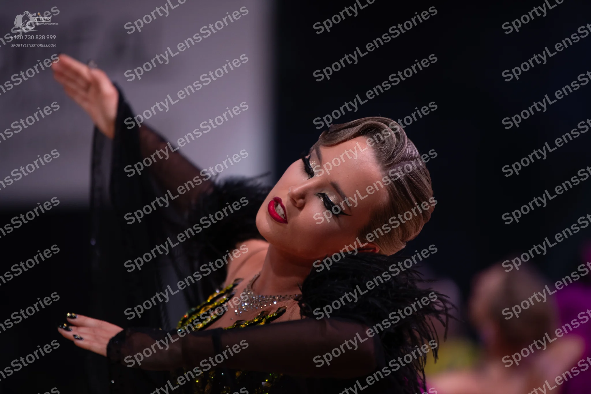 Dancesport competitor: Female dancer in foreground: black Latin-style dress with sheer long sleeves, bl