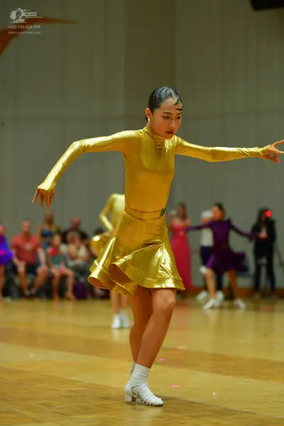 Dancesport competitor: Female dancer in foreground: gold metallic long-sleeved latin dress with a flare