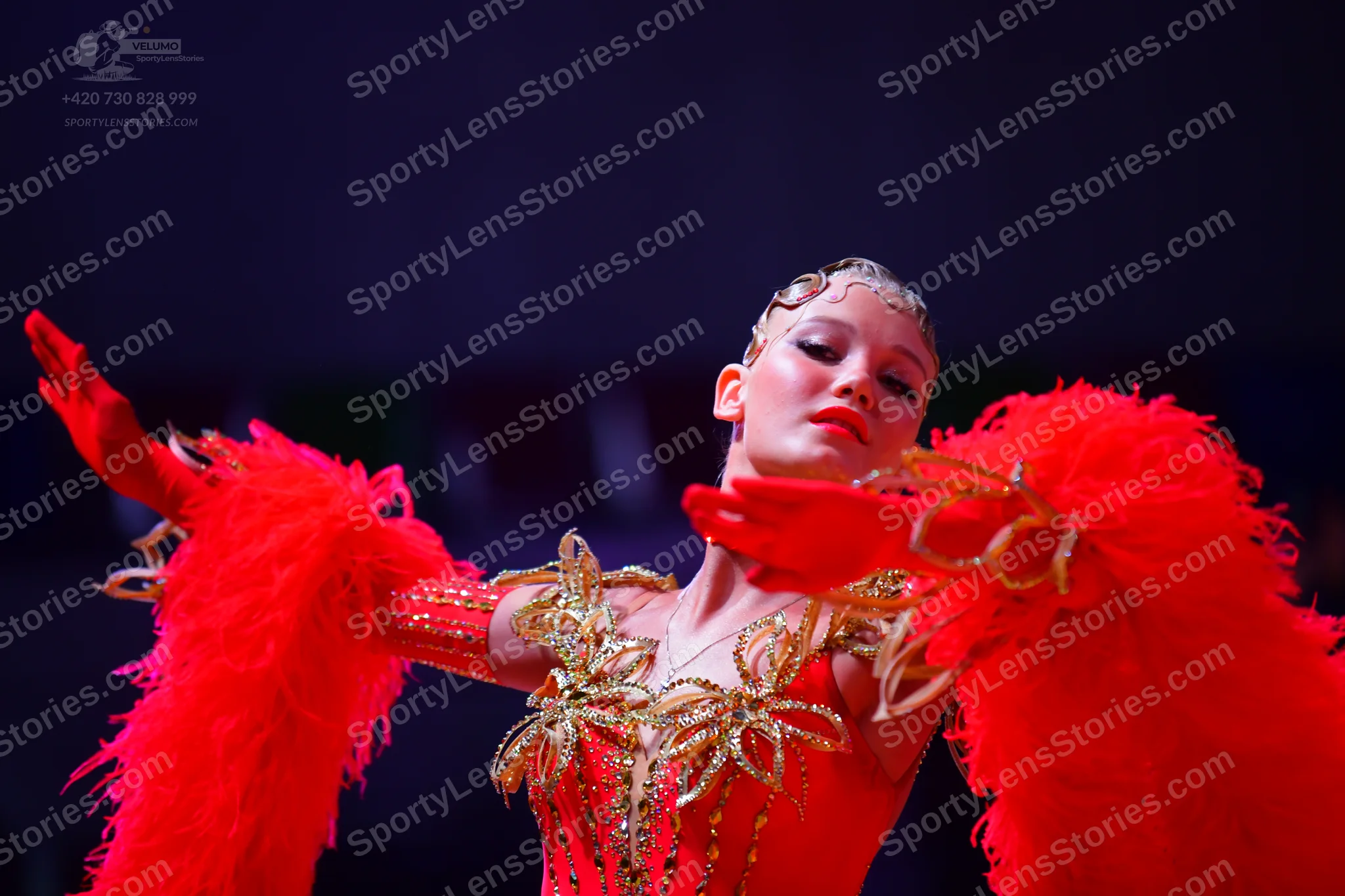 Dancesport competitor: Female dancer: red Latin dress with large red feather sleeves, gold metallic flo