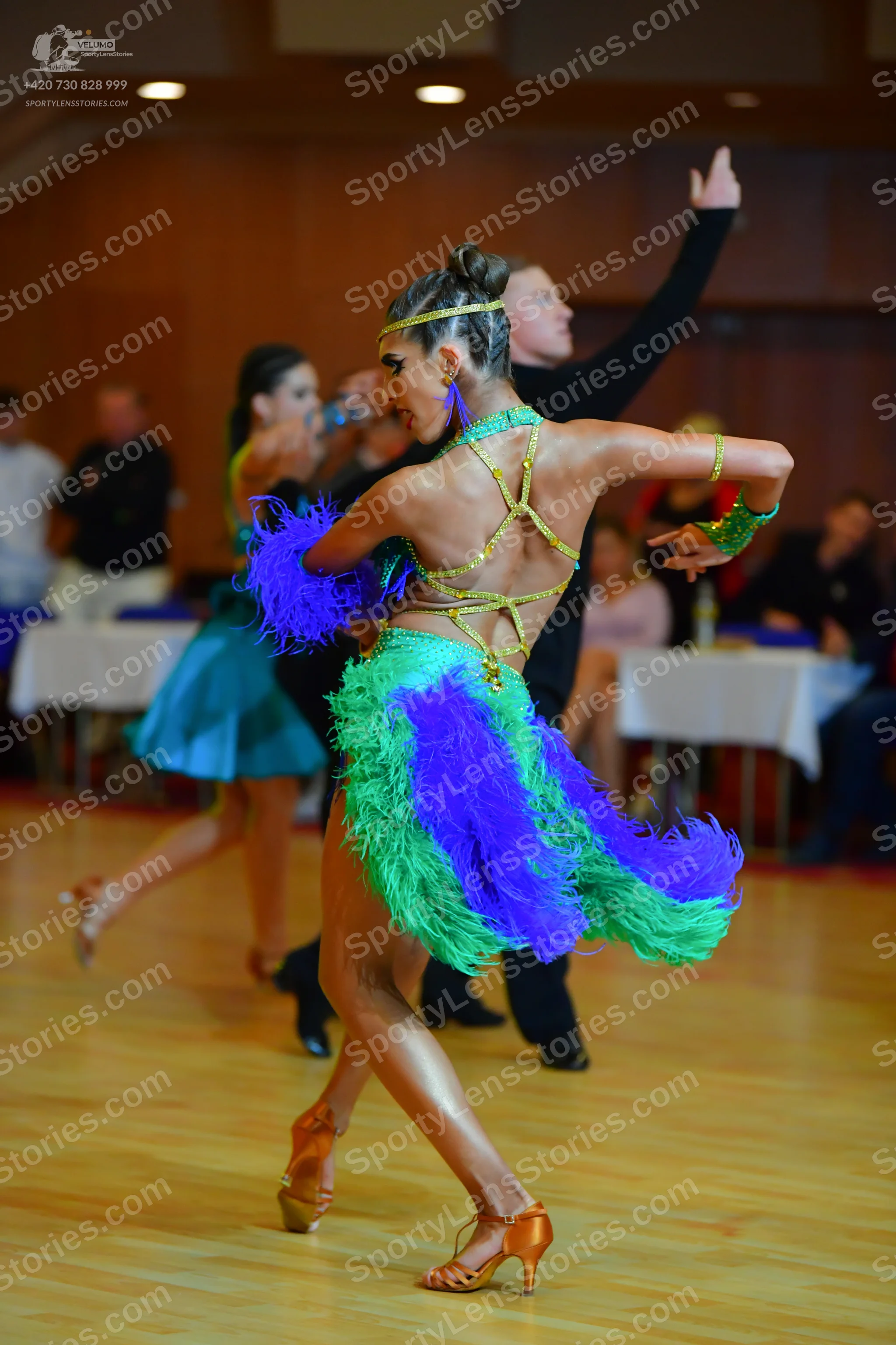 Dancesport competitor: Female dancer in foreground: green, blue, and purple feathered Latin dance dress