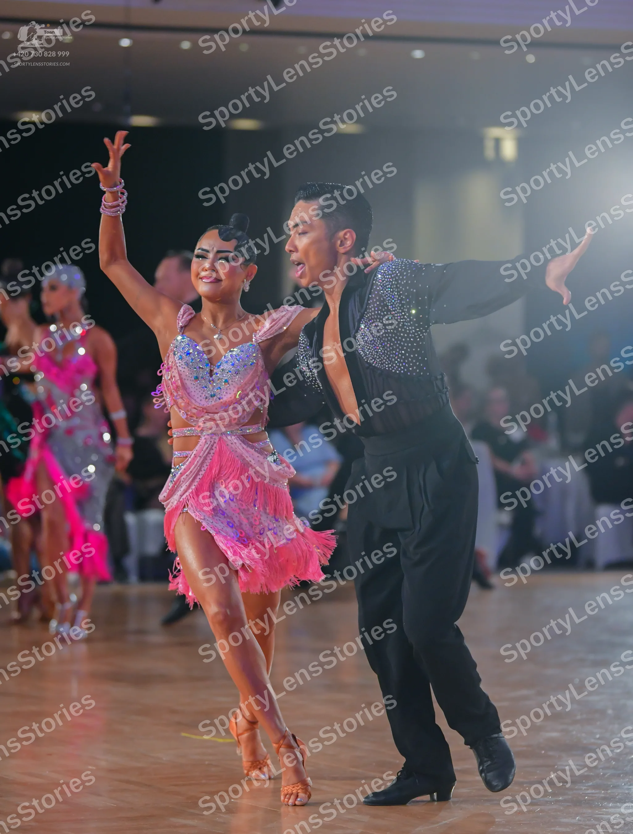 Dancesport competitor: Foreground female dancer: pink latin dance dress with sequins and fringe, and pi