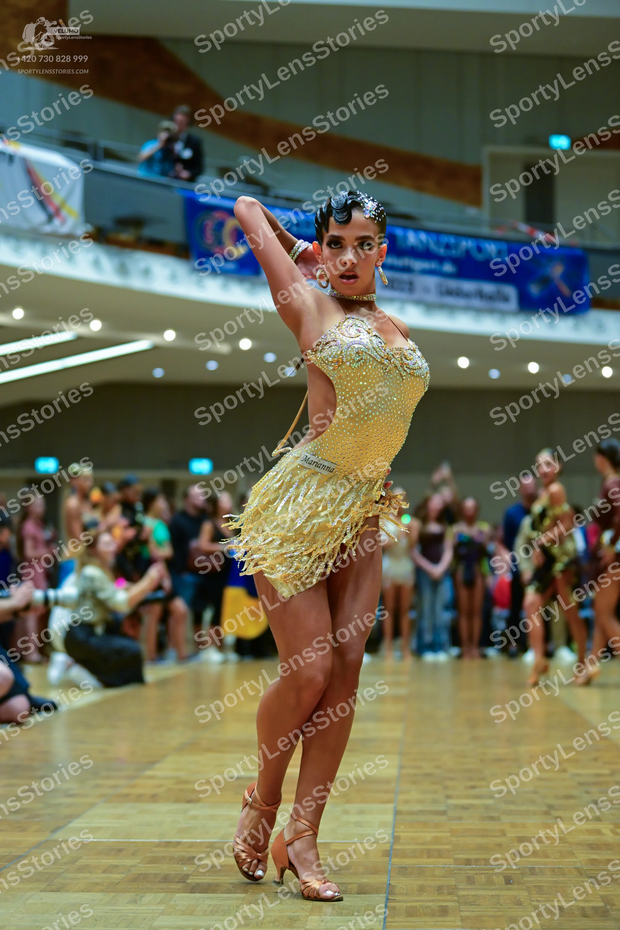 Dancesport competitor: Female dancer in foreground: gold Latin dance dress with sequins and fringe, and