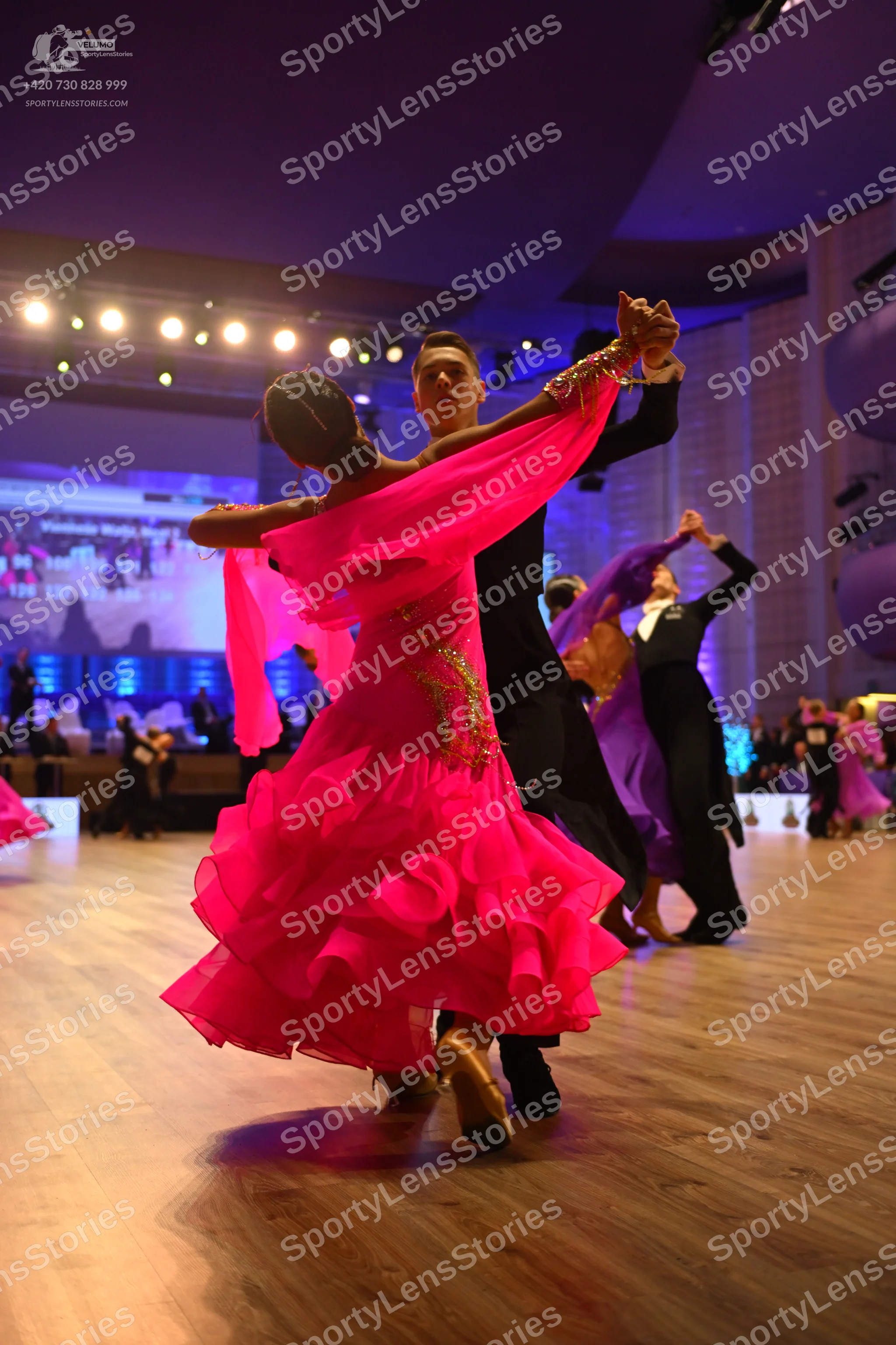 Dancesport competitor: Foreground female dancer: bright pink Latin dance dress with a layered, ruffled
