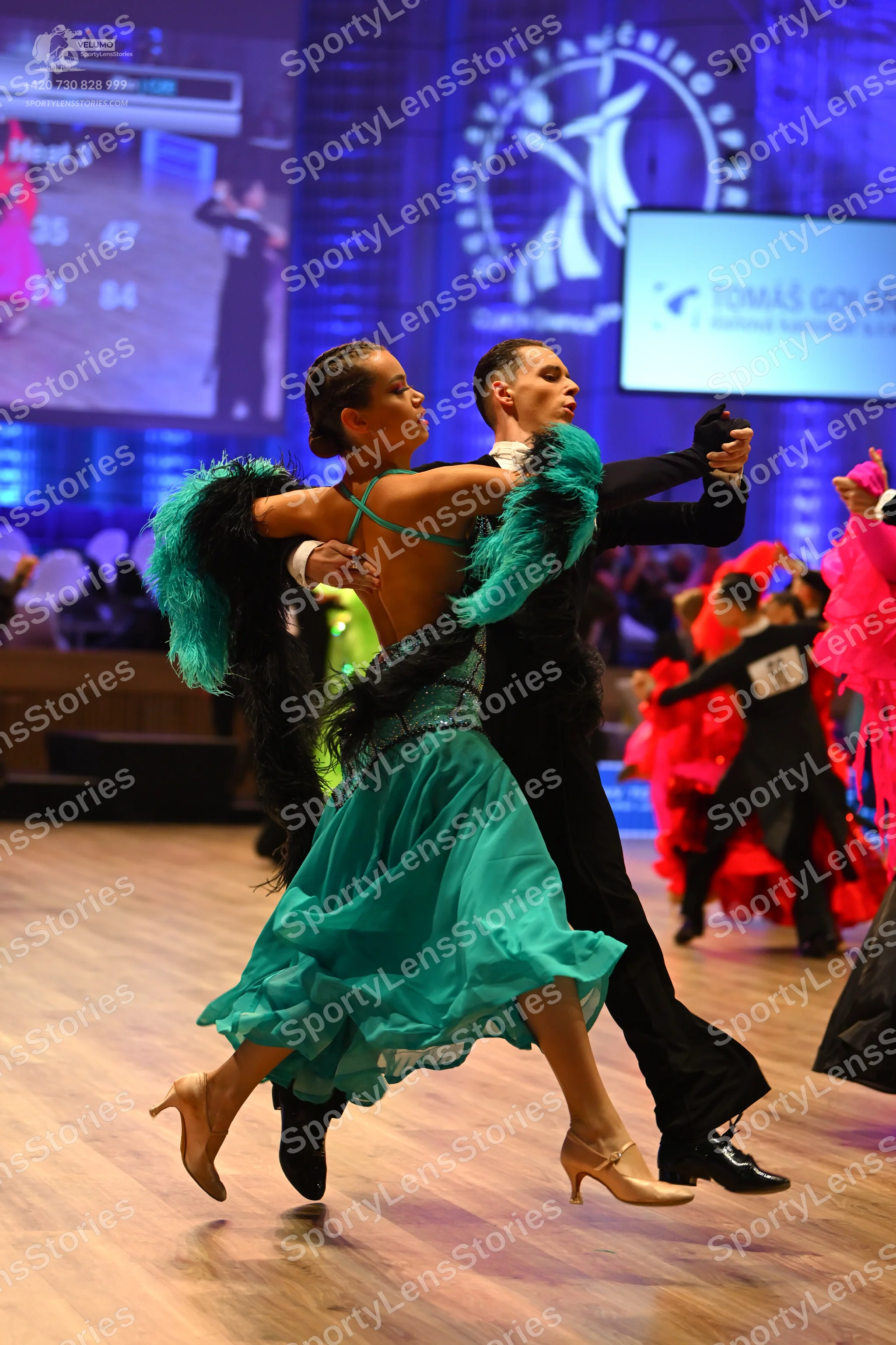 Dancesport competitor: Main female dancer: teal Latin dress with black feather-like trim on the arms, a
