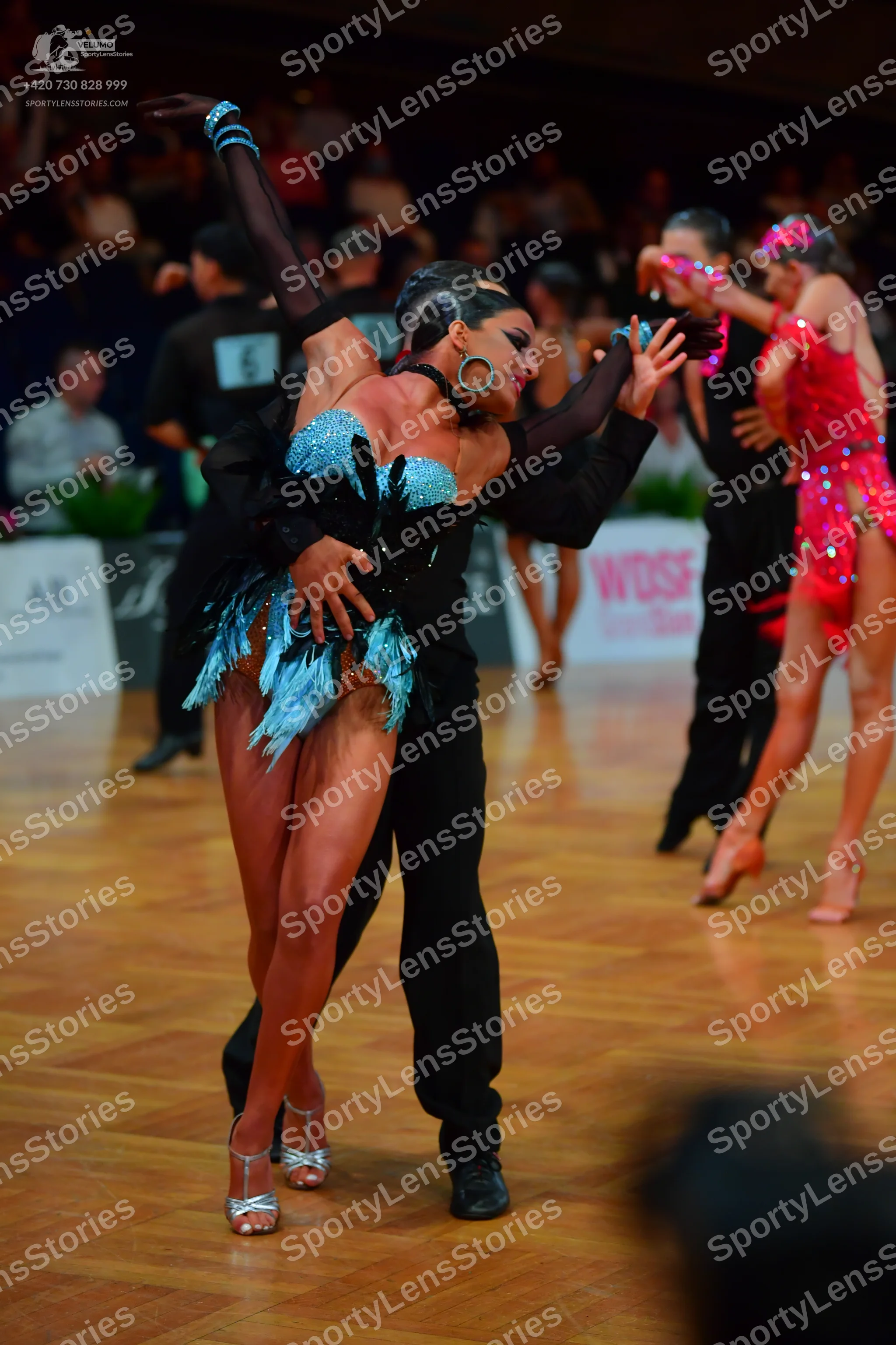Dancesport competitor: Foreground female dancer: teal blue sequin Latin dress with fringe and large hoo