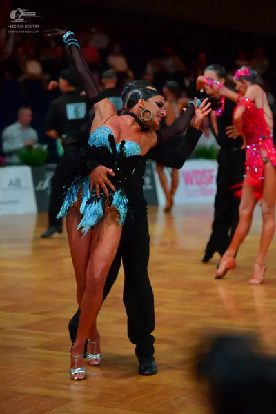 Dancesport competitor: Foreground female dancer: teal blue sequin Latin dress with fringe and large hoo