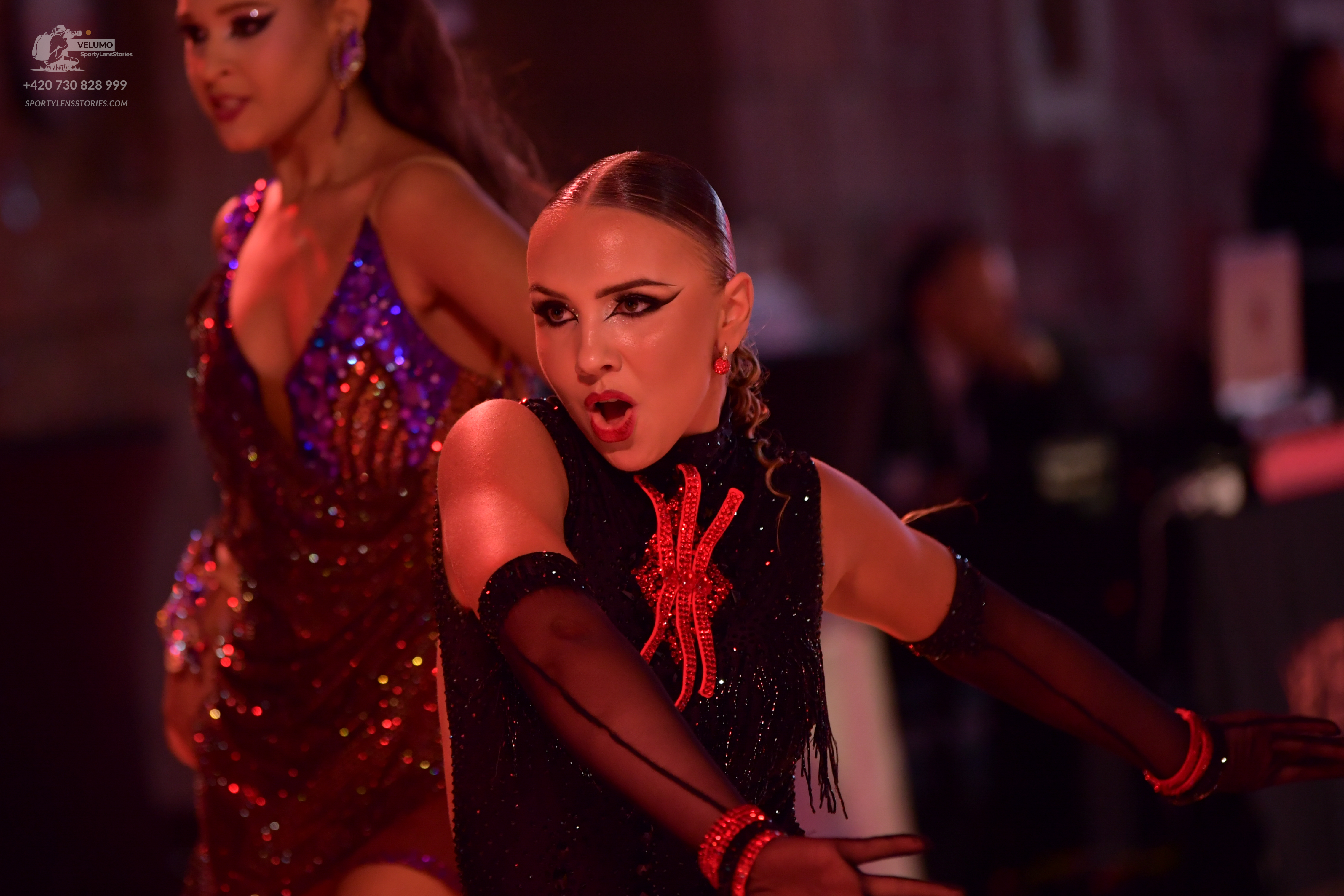 Dancesport competitor: Female dancer in foreground: black latin dress with a red and gold decorative mo