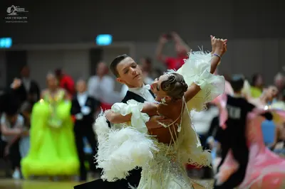 Dancesport competitor: Foreground female dancer: cream-colored ballroom dress with large, voluminous ru