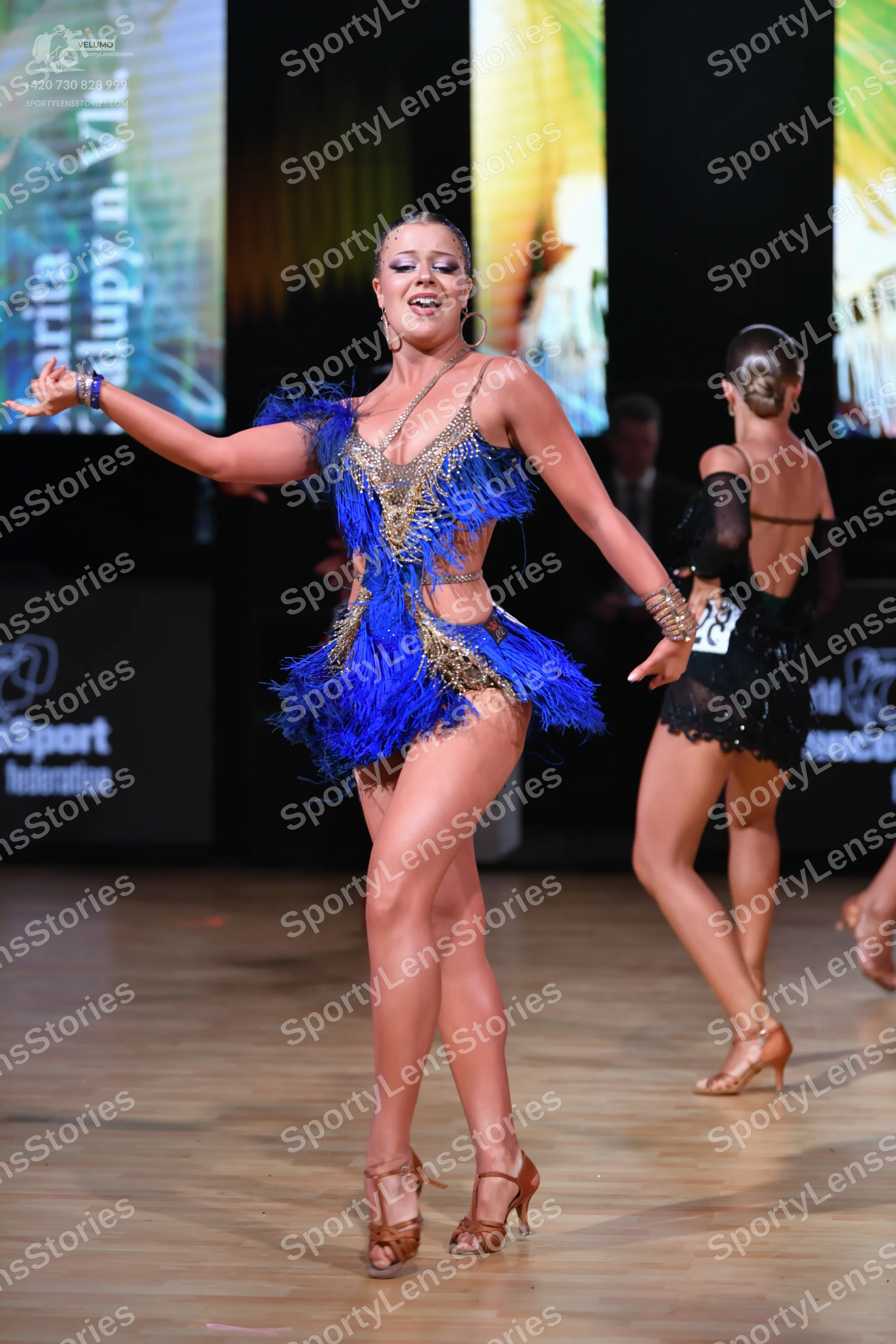Dancesport competitor: Female dancer in foreground: blue Latin dress with gold sequins and blue fringe