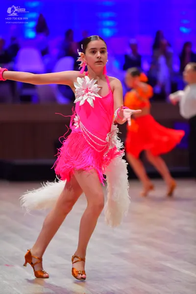 Dancesport competitor: Female dancer in foreground: bright pink latin dance dress with fringe, large wh