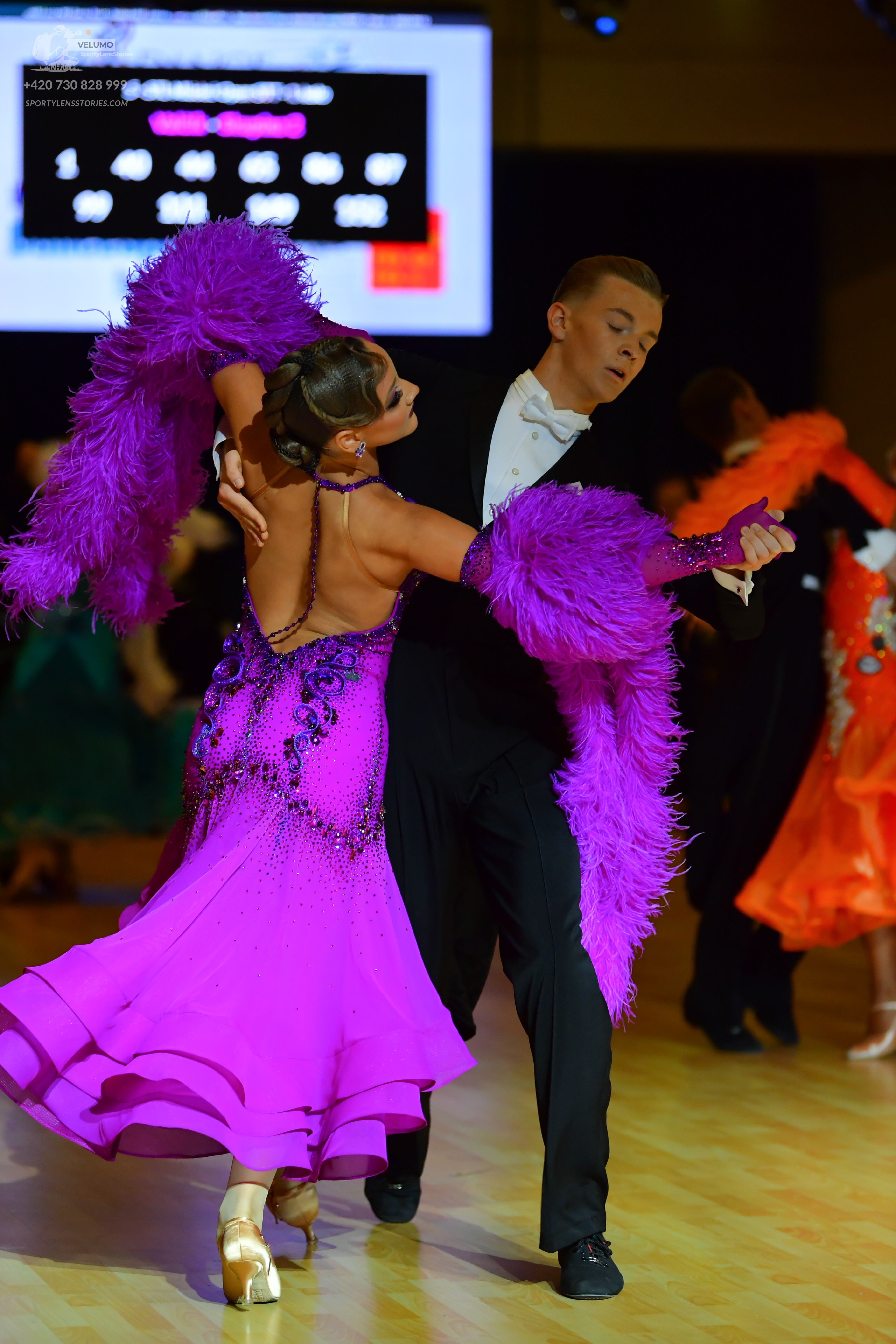 Dancesport competitor: Female dancer in foreground: magenta/purple dress with a sequined bodice and lar