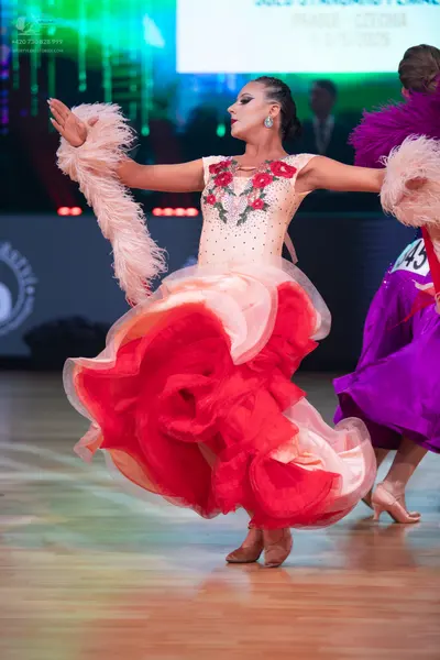 Dancesport competitor: Female dancer (foreground): cream-colored ballroom dress with red floral embroid