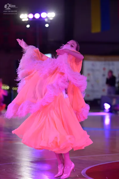Dancesport competitor: Female dancer: voluminous, neon pink/coral ballroom dress with many layers of fe
