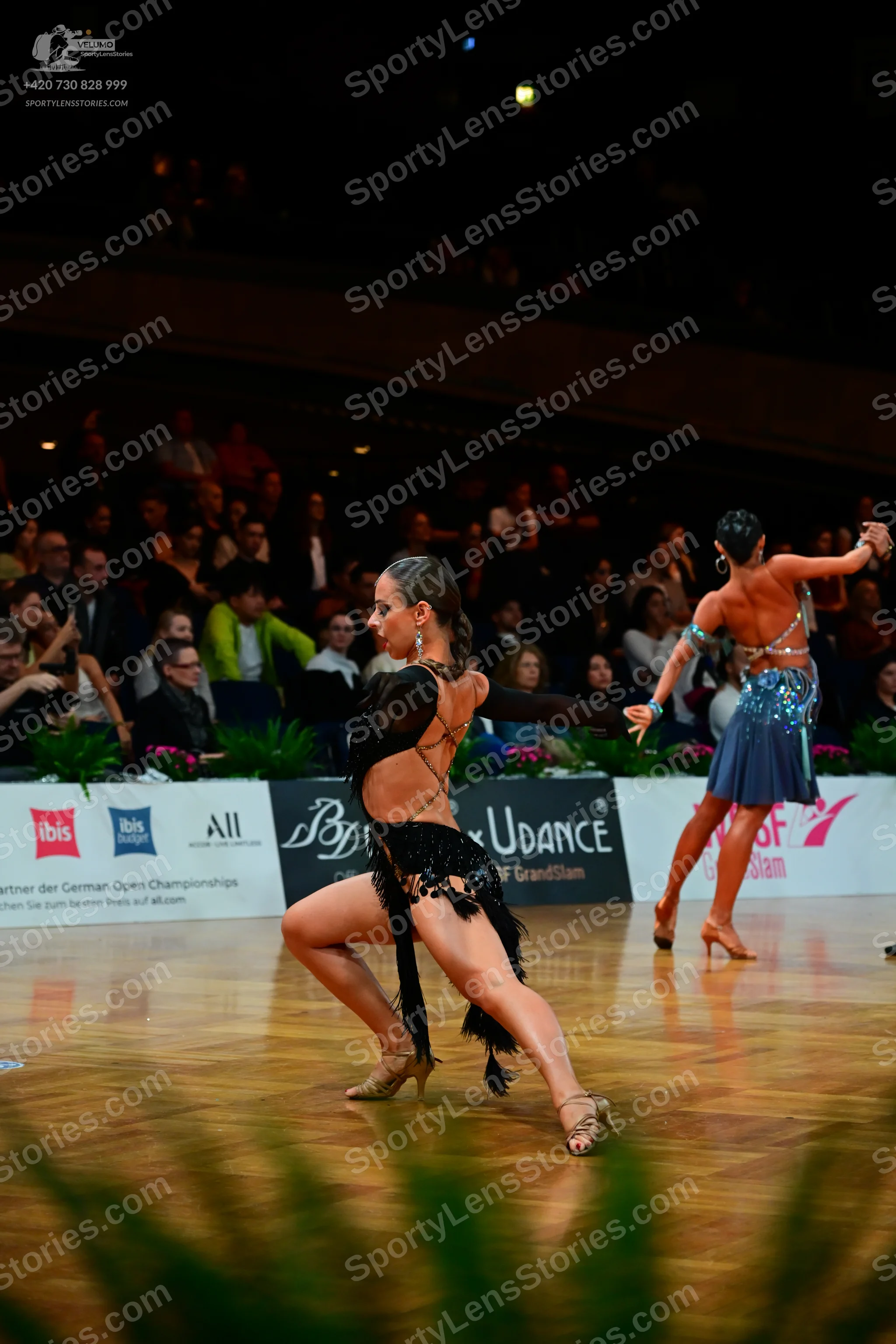 Dancesport competitor: Female dancer in foreground: black latin dress with sheer panels and fringe