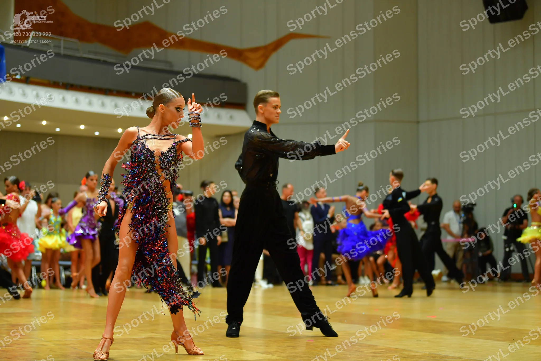 Dancesport competitor: Foreground female dancer: dark Latin dress with multicolored sequins (pink, blue
