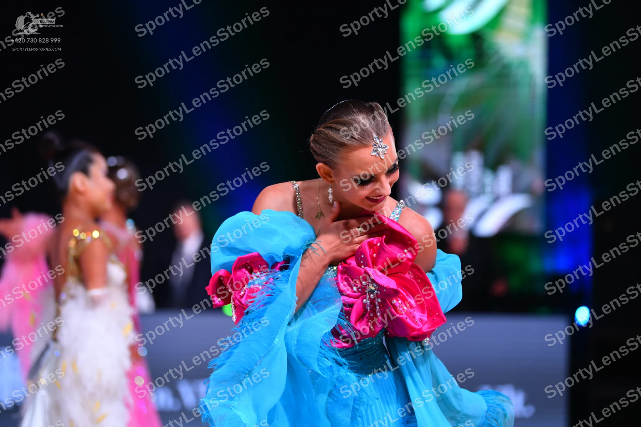 Dancesport competitor: Female dancer in foreground: bright blue voluminous ballroom dress with large pi