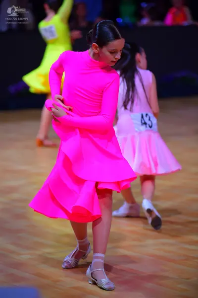 Dancesport competitor: Female dancer in foreground: bright pink, long-sleeved dress with a layered, ruf