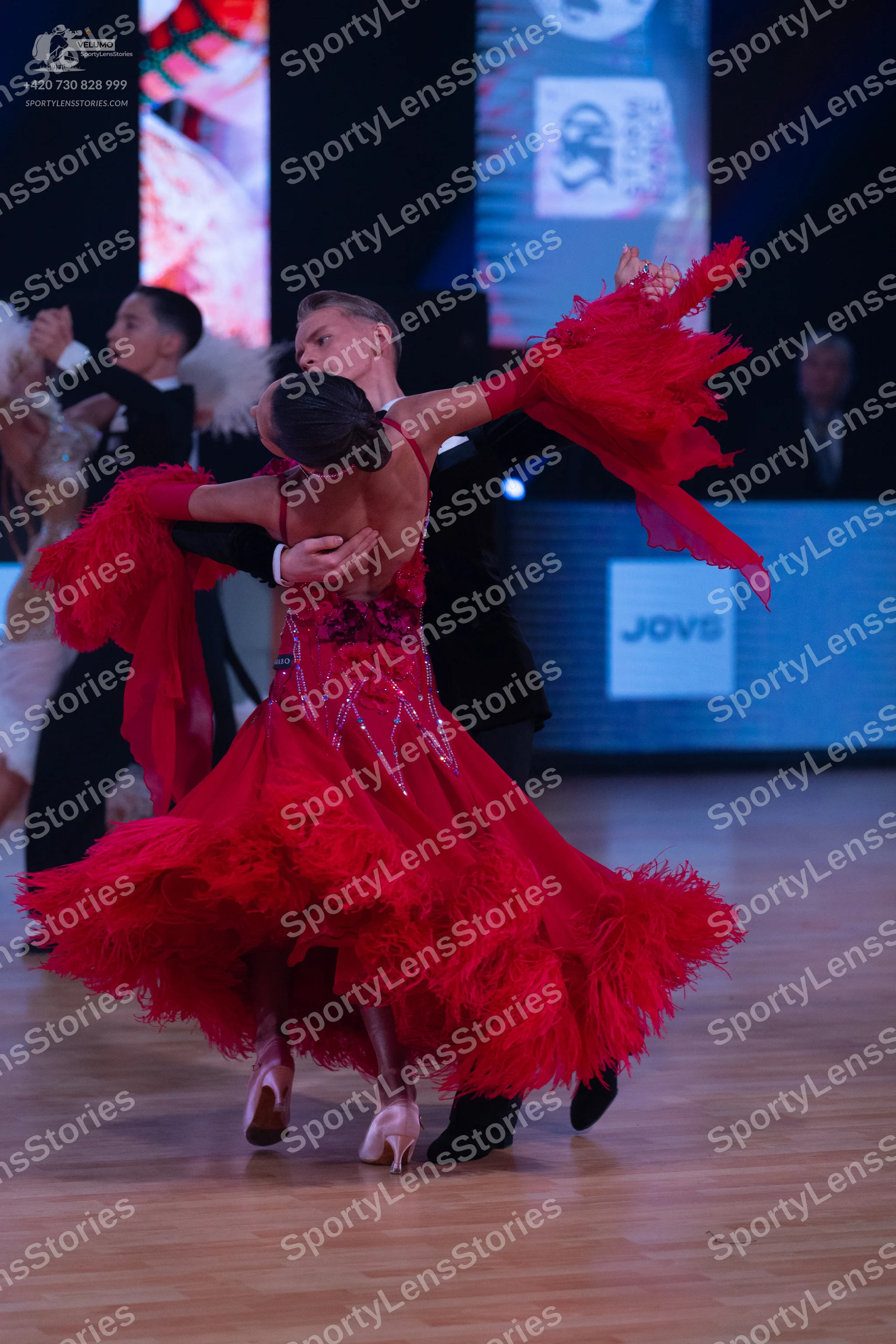 Dancesport competitor: Female dancer in foreground: red Latin dress with sequins on the bodice and a vo