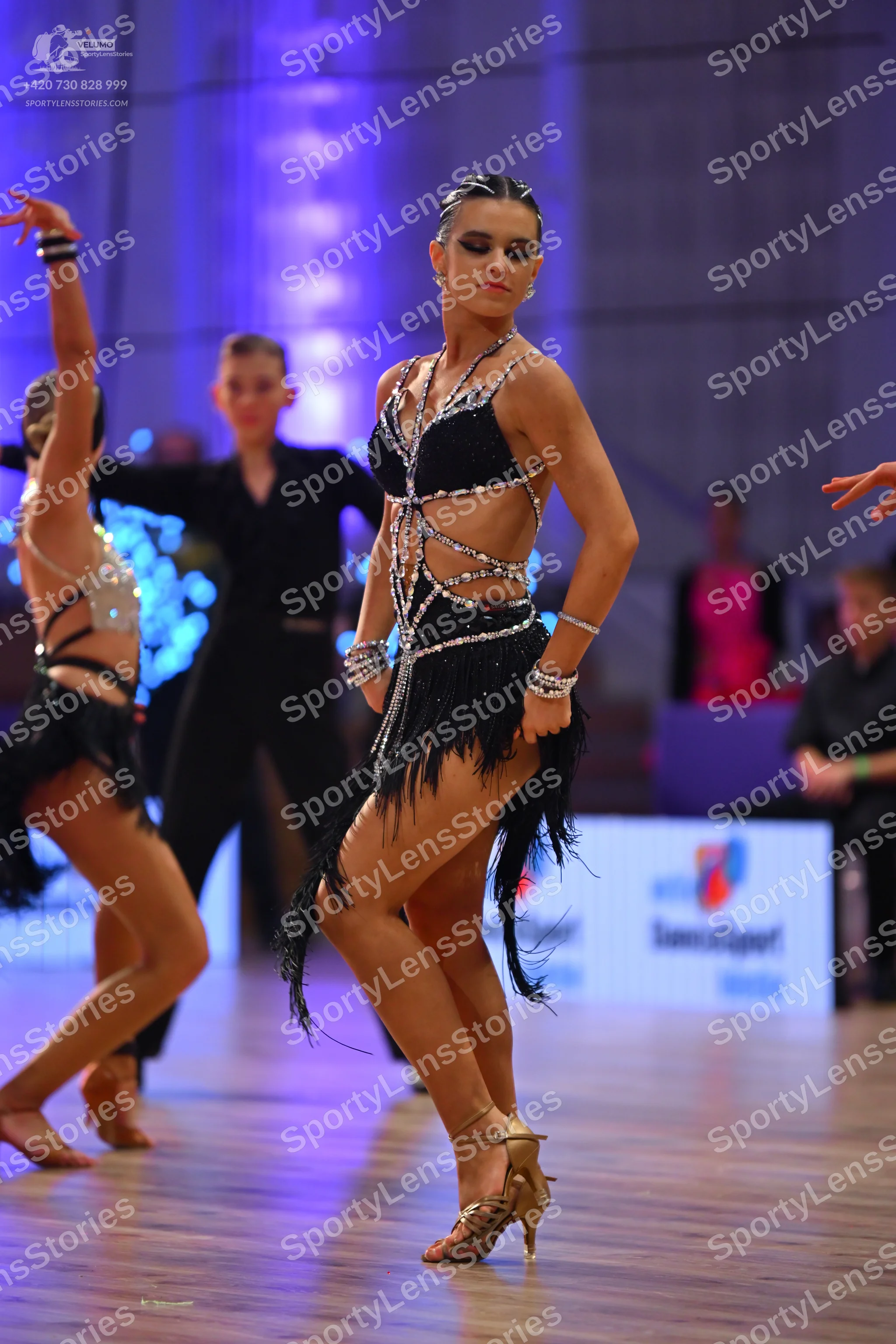 Dancesport competitor: Female dancer in foreground: black Latin dress with silver/white rhinestone-embe