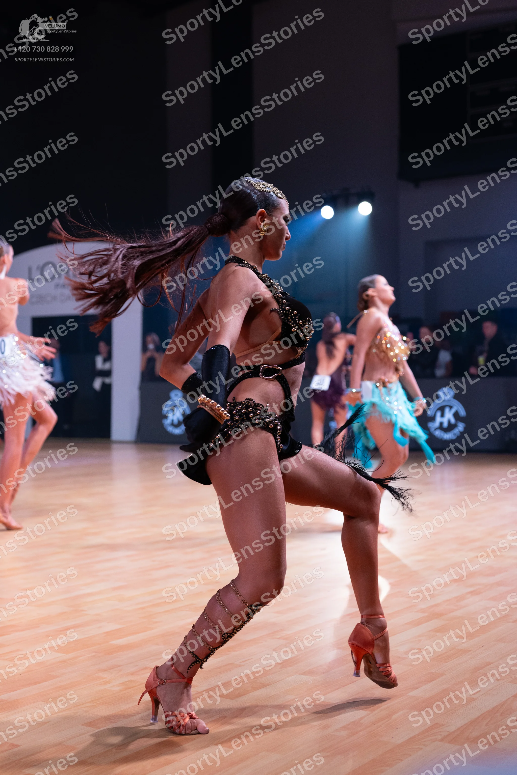 Dancesport competitor: Female dancer in foreground: black Latin dance costume heavily embellished with