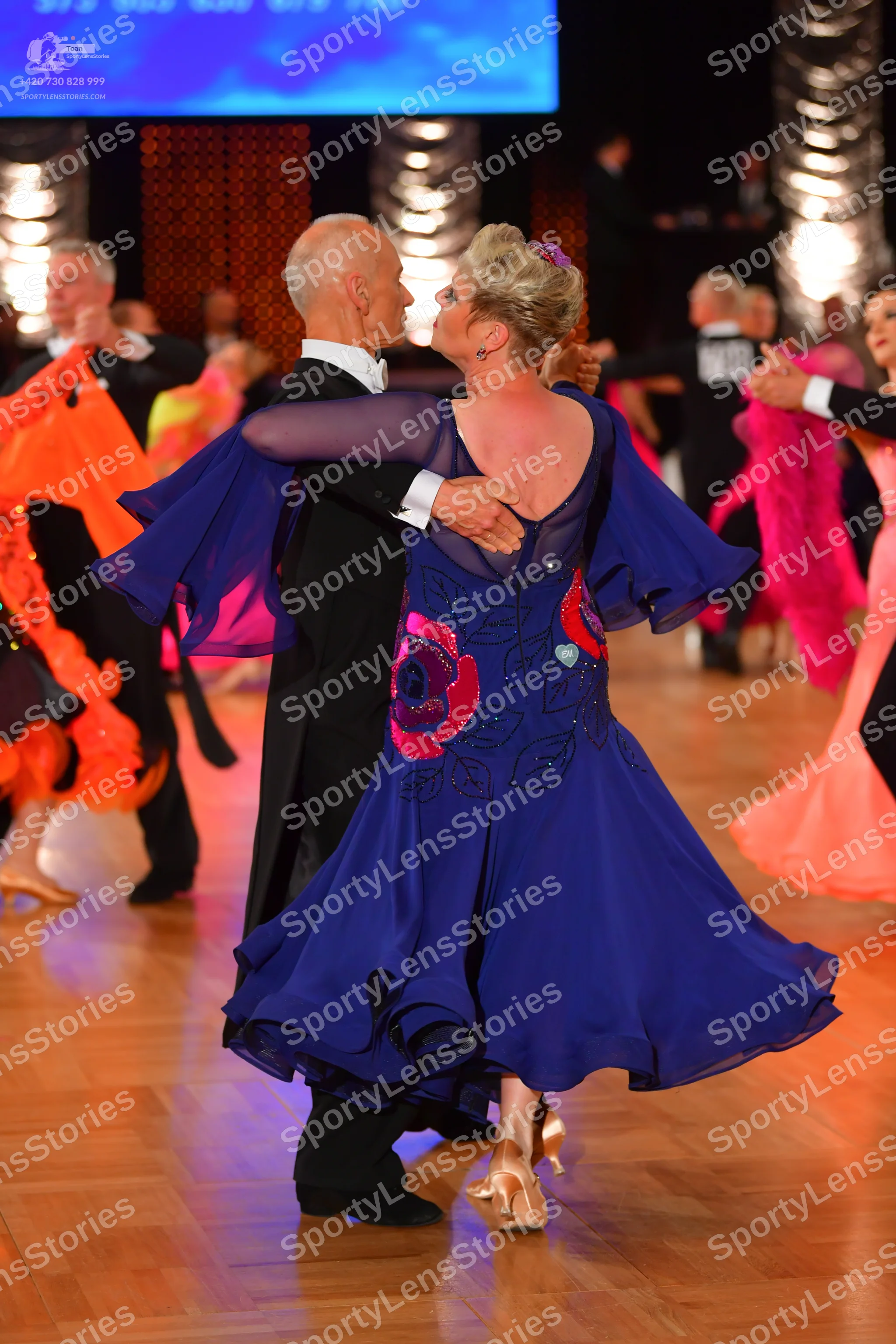 Dancesport competitor: Foreground female dancer: deep purple/blue flowing dress with a large red floral