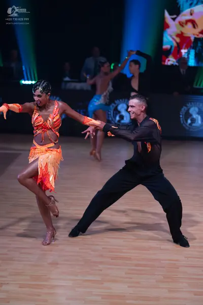 Dancesport competitor: Foreground female dancer: orange and yellow Latin dance dress with a fringed ski