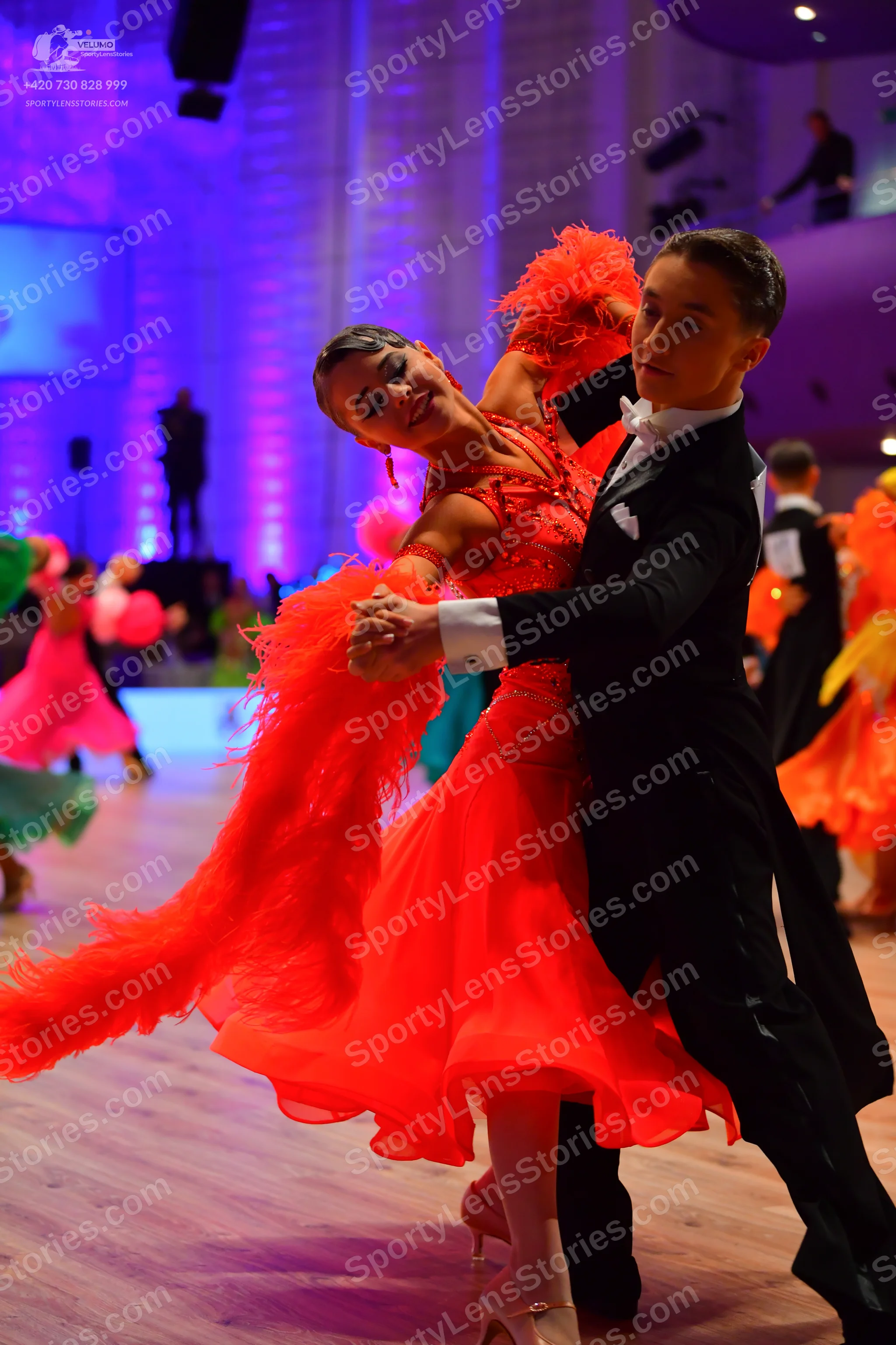 Dancesport competitor: Female dancer in foreground: bright red latin dress with large red feather/fring