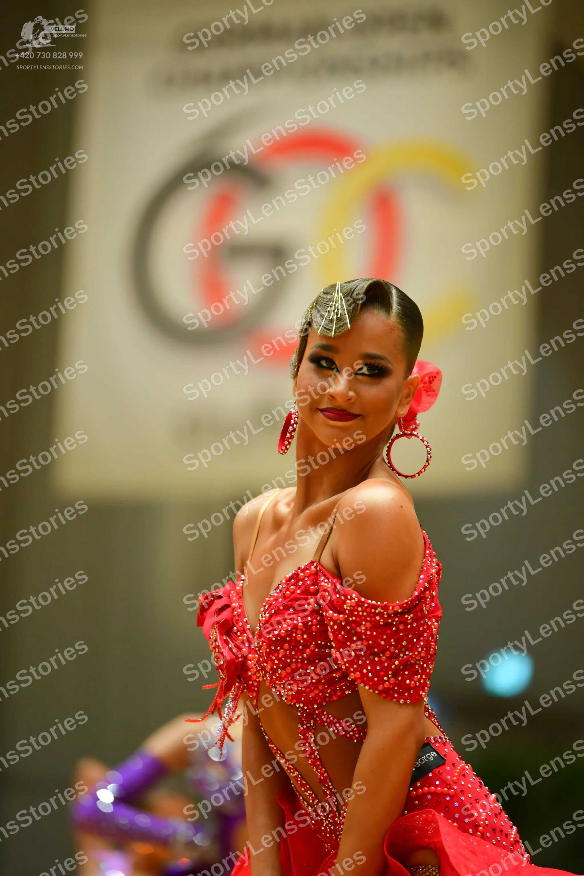 Dancesport competitor: Female dancer in foreground: red Latin dance dress with rhinestones and cutouts,