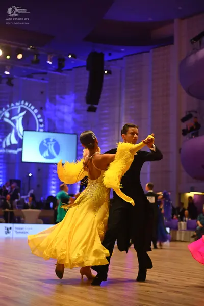 Dancesport competitor: Female dancer: yellow ballgown with feather trim on the sleeves and sequin embel