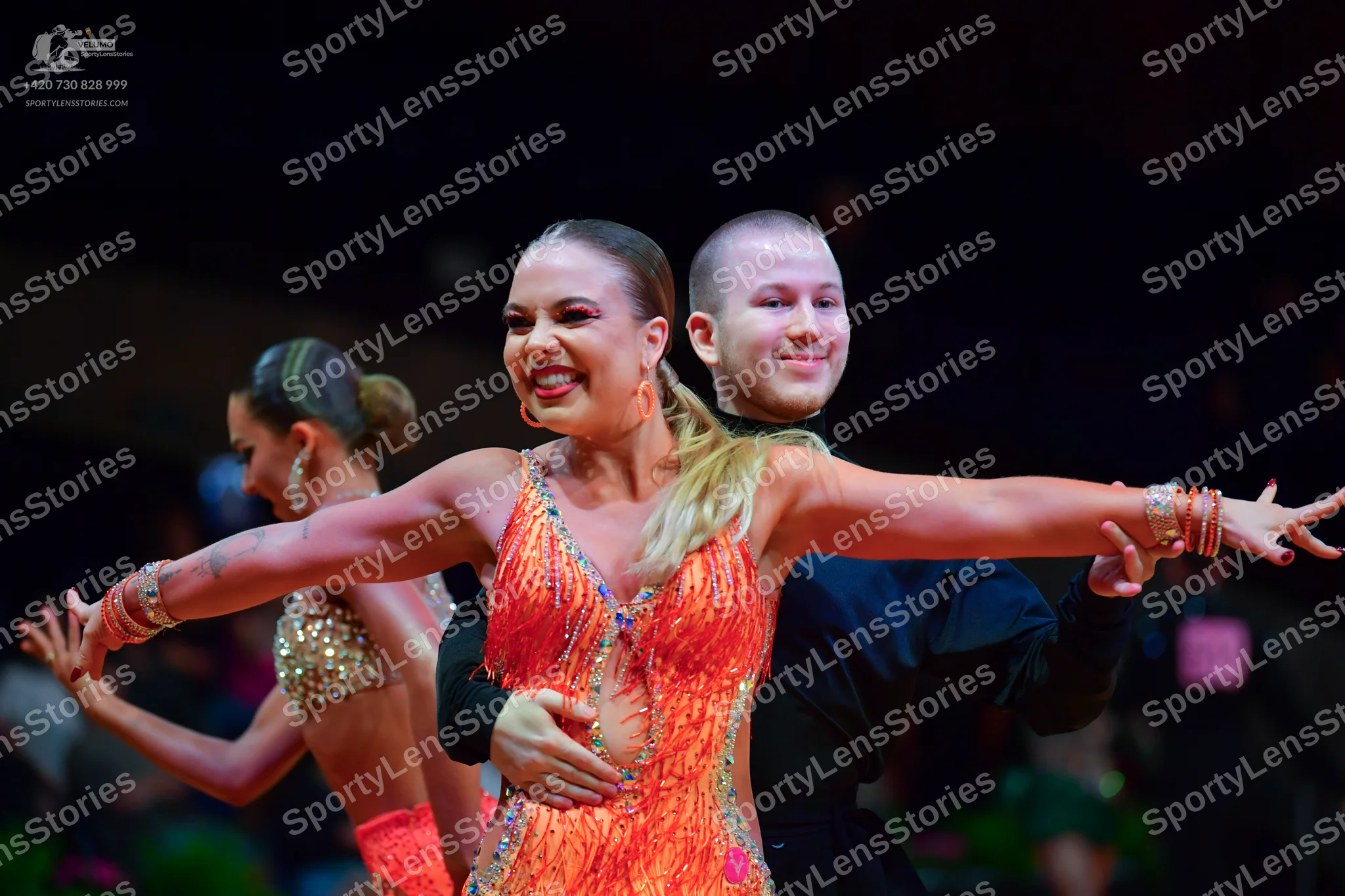 Dancesport competitor: Central female dancer: orange/coral Latin dress heavily embellished with silver