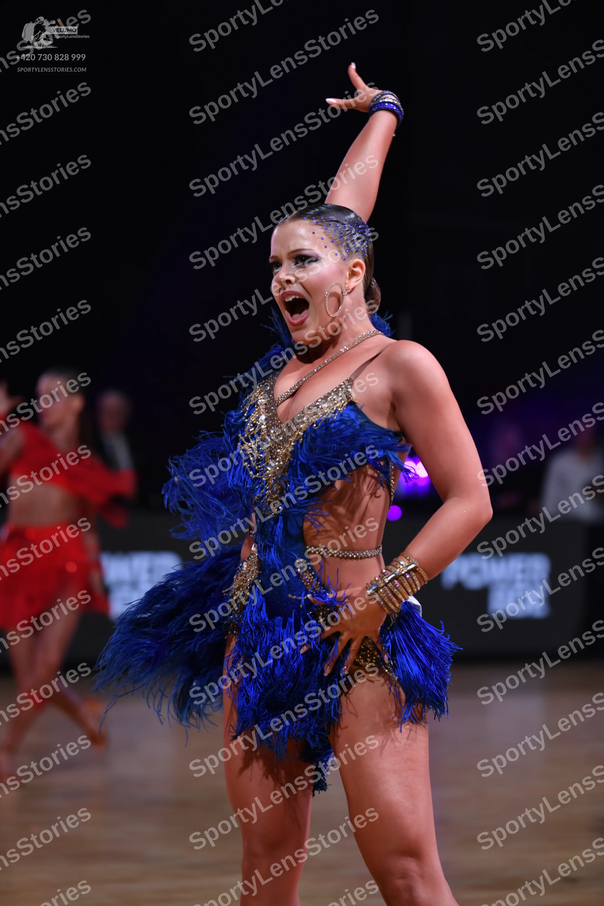 Dancesport competitor: Female dancer in foreground: blue latin costume with blue feathers, gold/silver