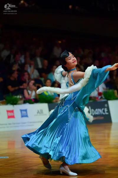 Dancesport competitor: Female dancer: teal-blue ballroom gown with a pleated skirt, featuring white fau