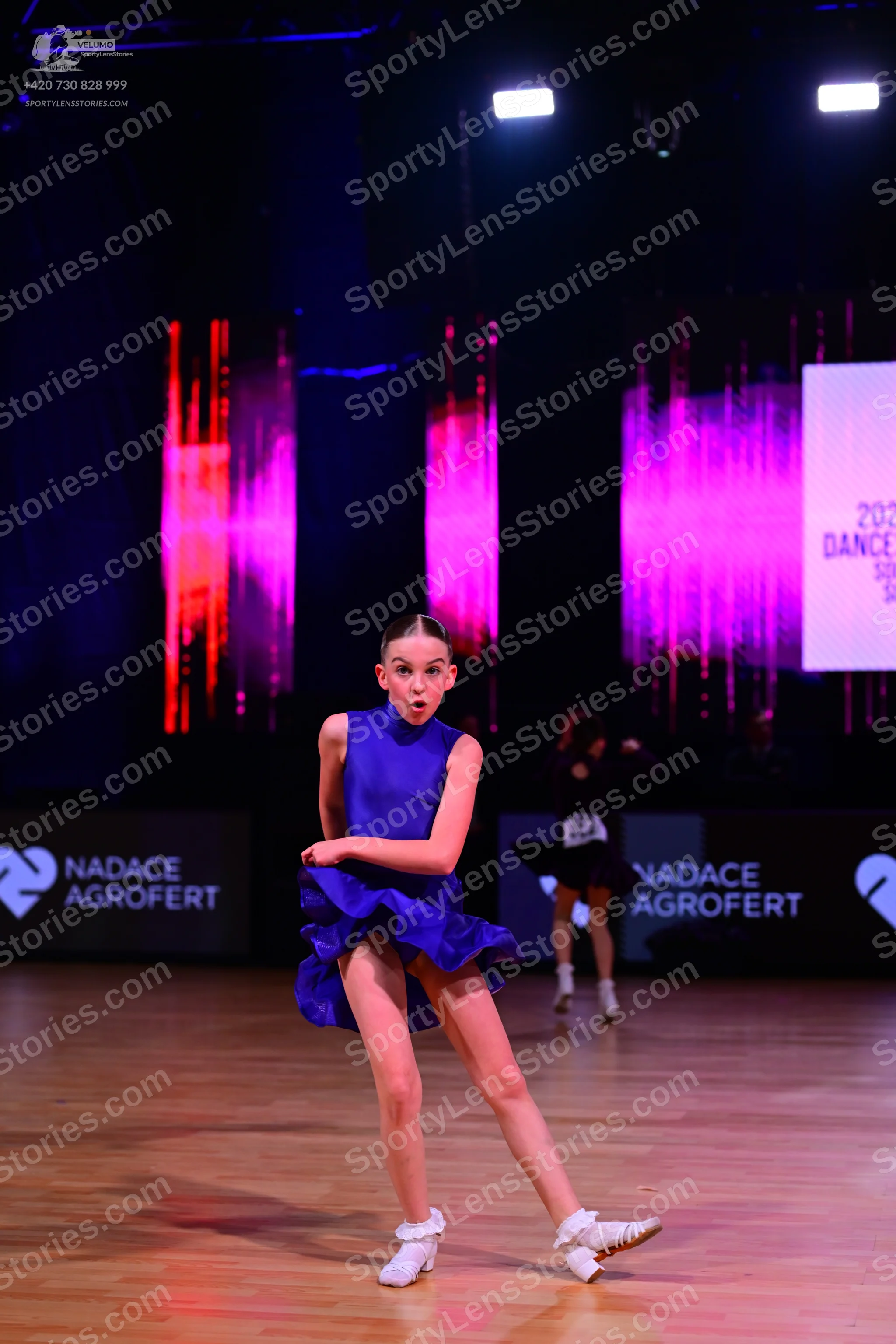 Dancesport competitor: Female dancer in foreground: blue, sleeveless, short latin dress with a ruffled 