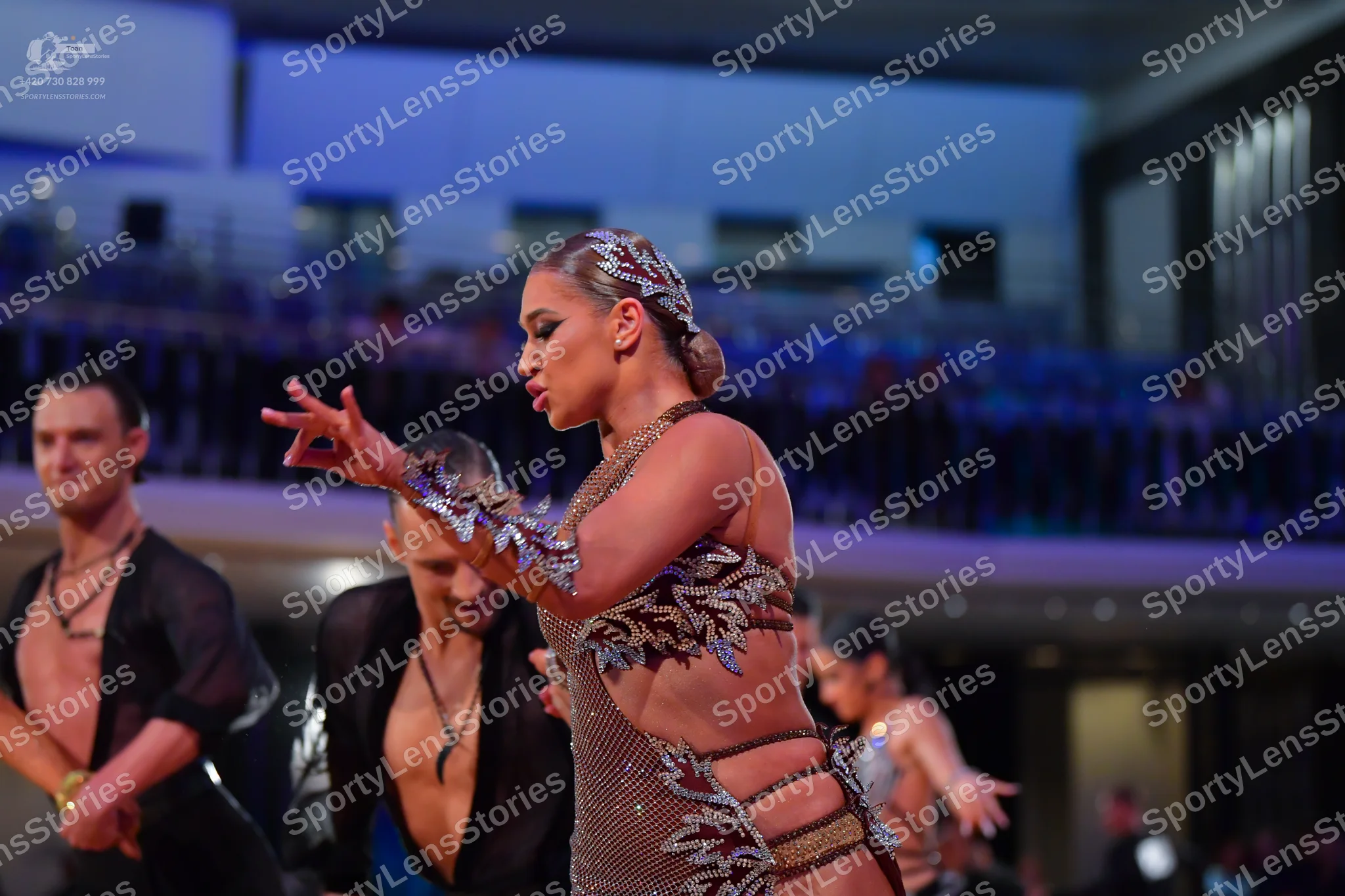 Dancesport competitor: Foreground female dancer: tan/nude Latin dance costume featuring intricate dark,