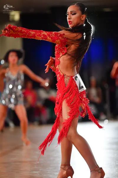 Dancesport competitor: Female dancer in foreground: red Latin dress with lace, sequins, and long red fr