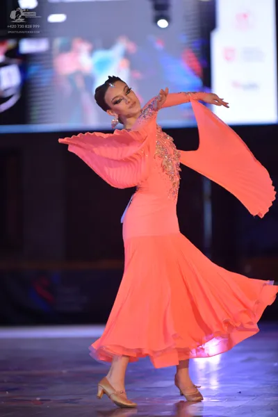 Dancesport competitor: Female dancer: peach-colored long flowing dress with wide sleeves and sequin/cry