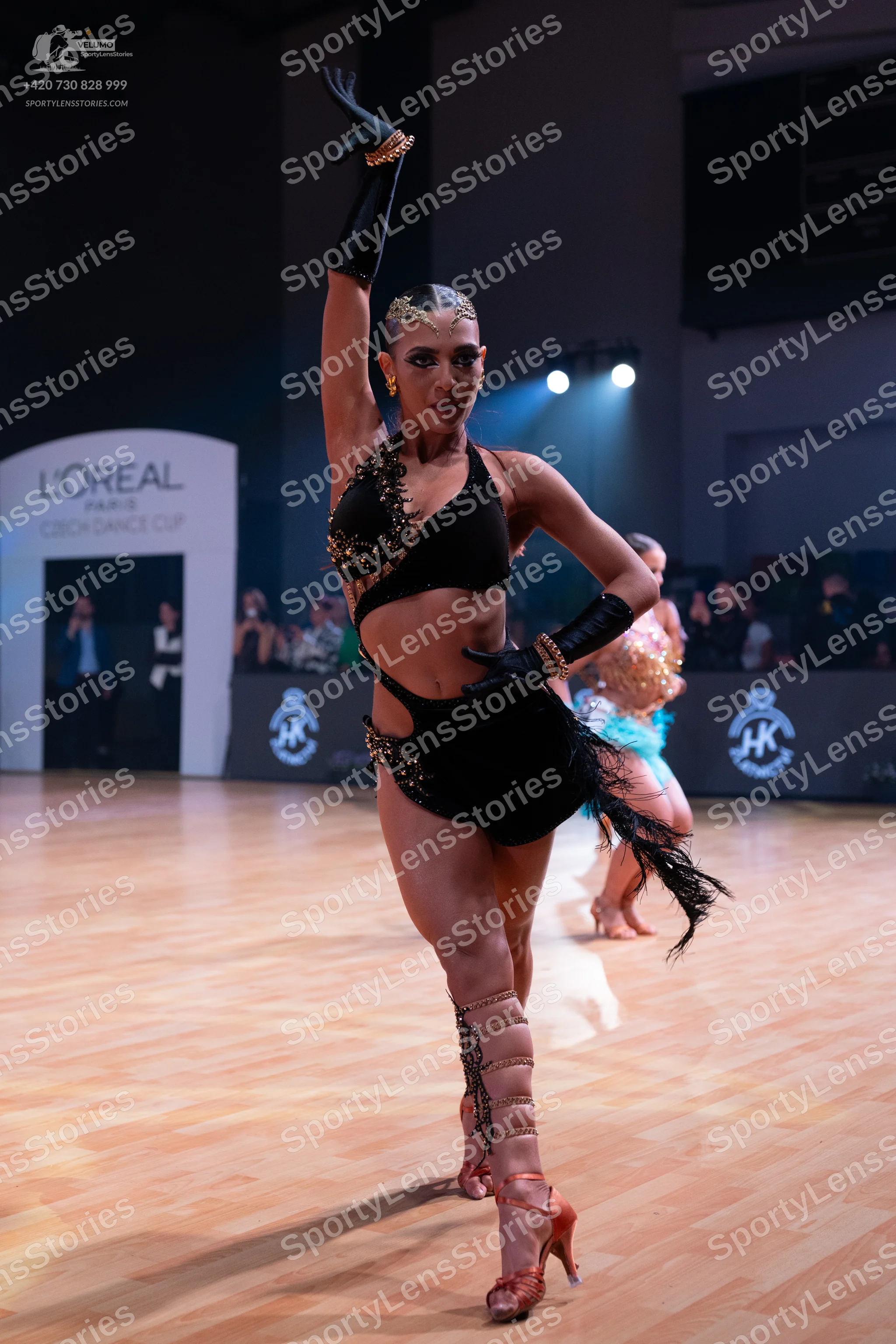 Dancesport competitor: Female dancer in foreground: black latin dance costume with gold sequin embellis