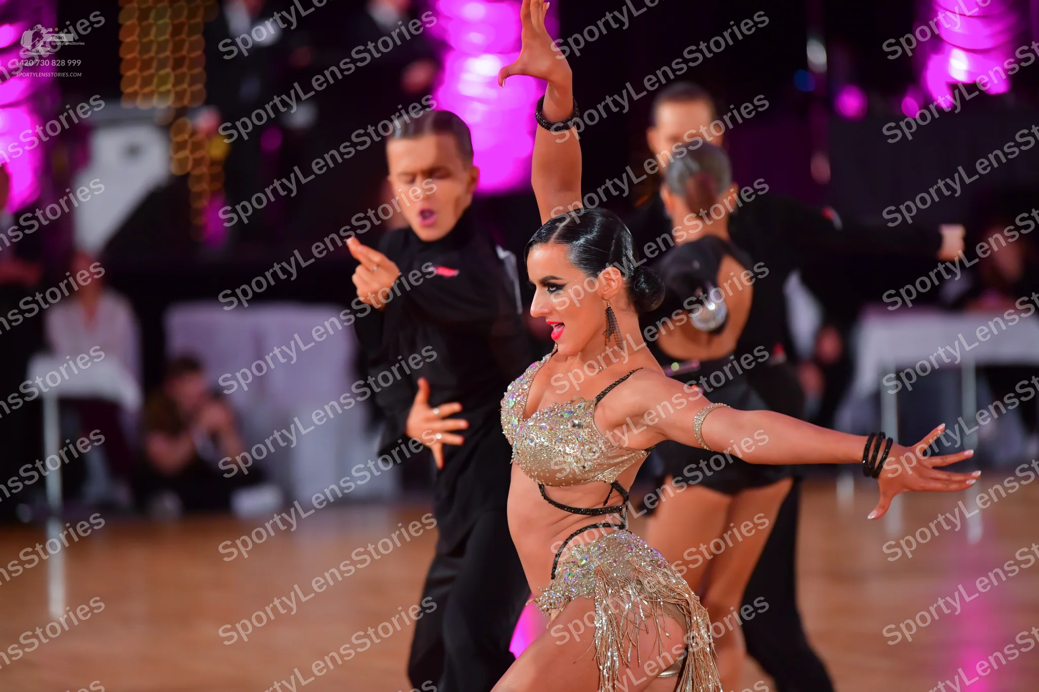 Dancesport competitor: Foreground female dancer: gold sequined Latin dance costume with thin black stra
