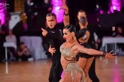 Dancesport competitor: Foreground female dancer: gold sequined Latin dance costume with thin black stra
