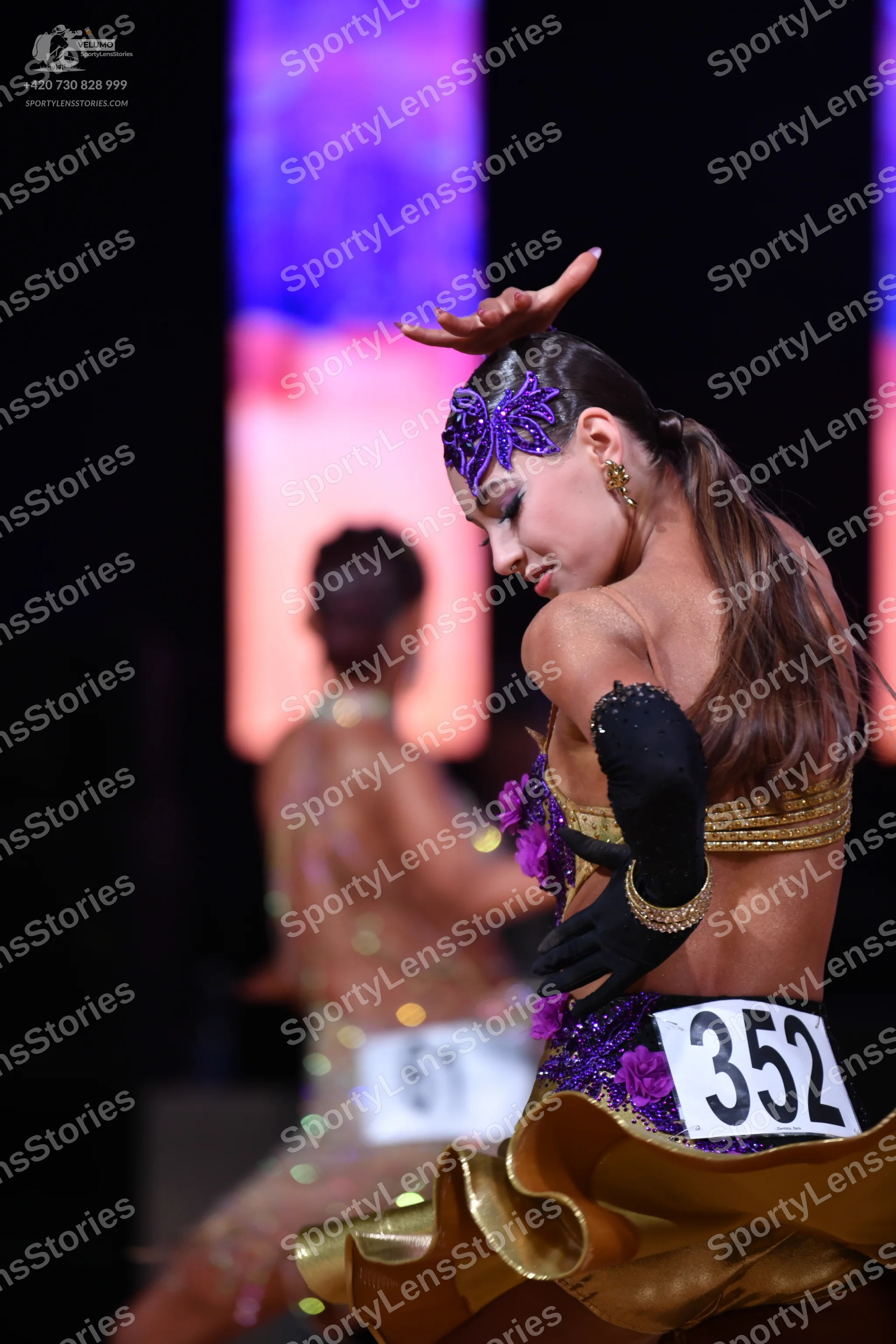 Dancesport competitor: Female dancer in foreground: purple and gold latin dress with floral embellishme