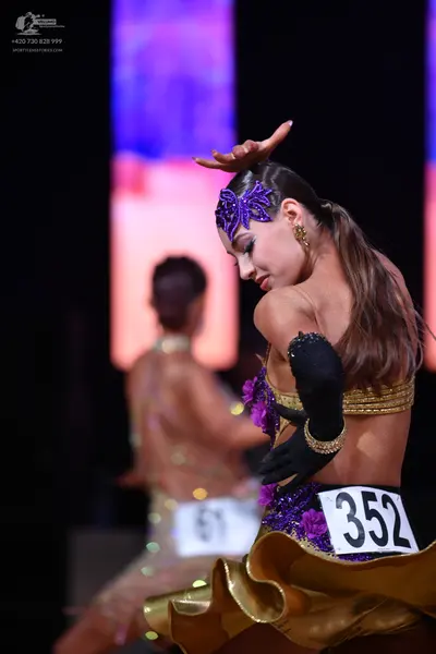 Dancesport competitor: Female dancer in foreground: purple and gold latin dress with floral embellishme