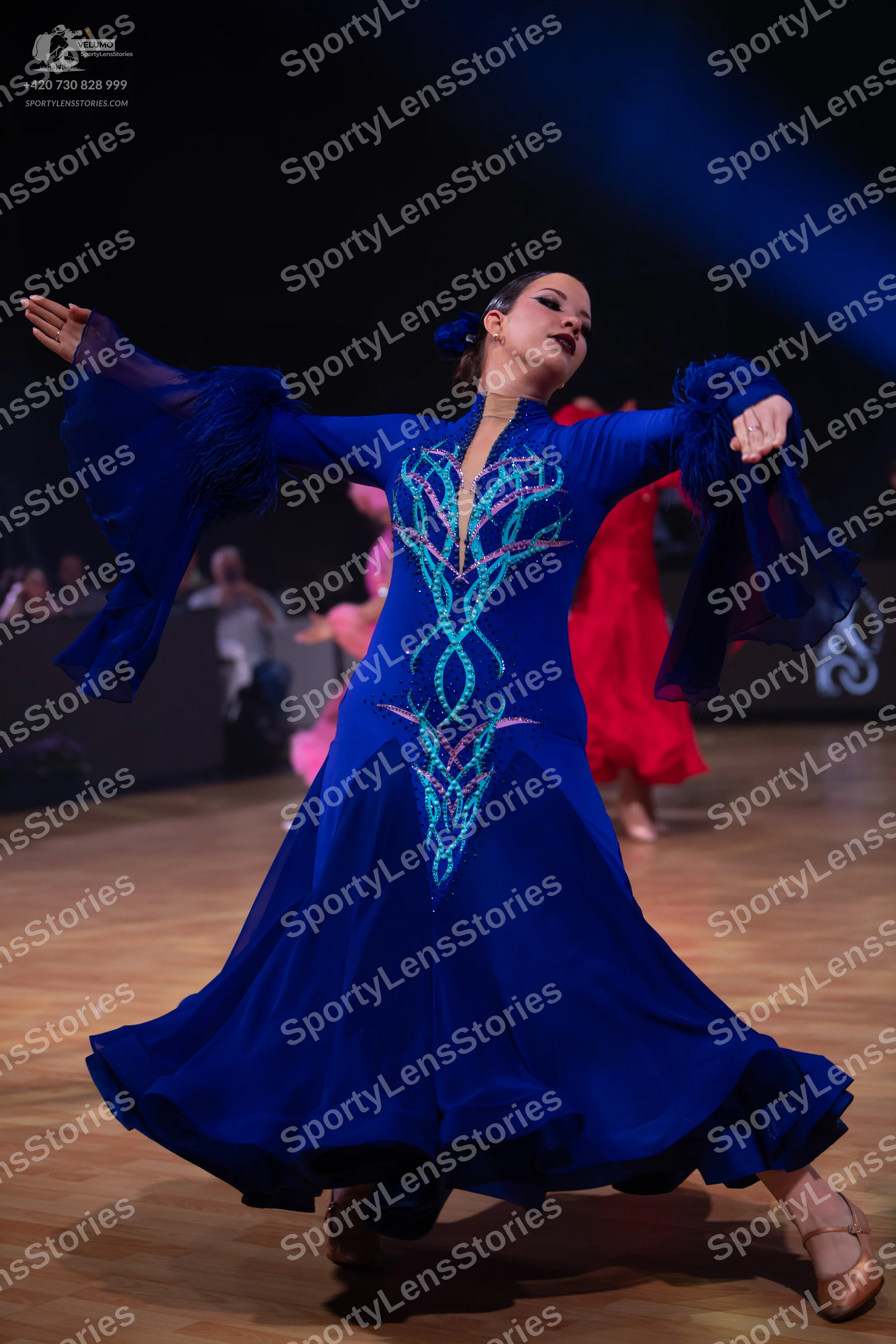 Dancesport competitor: Female dancer in foreground: blue long-sleeved ballroom dress with flared sleeve