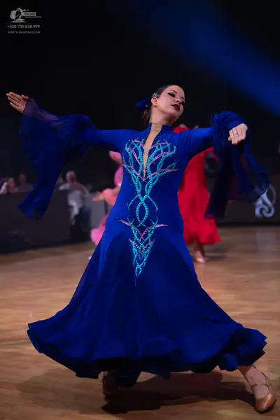 Dancesport competitor: Female dancer in foreground: blue long-sleeved ballroom dress with flared sleeve