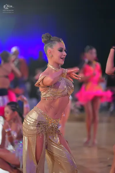Dancesport competitor: Female dancer in foreground: gold two-piece Latin costume consisting of a sequin