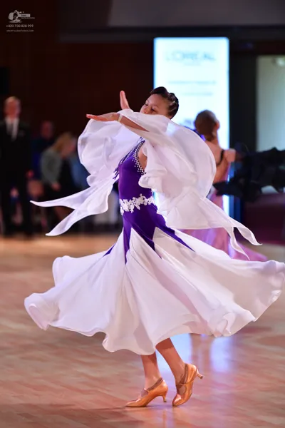 Dancesport competitor: Female dancer in foreground: white and purple ballroom dress with a large, flowi